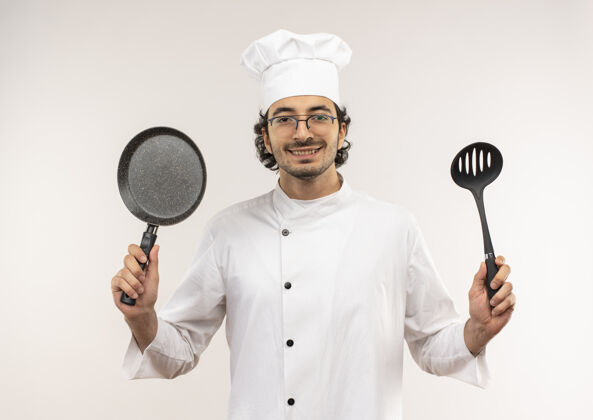 微笑面带微笑的年轻男厨师穿着厨师制服 戴着眼镜 手里拿着煎锅和抹刀 隔离在白墙上平底锅制服厨师
