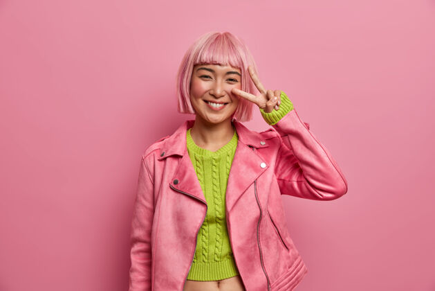 女士可爱的微笑着的亚洲年轻女子 粉色头发 用v字表示和平 用两个手指遮住眼睛室内象征年轻
