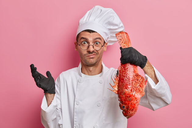 男人犹豫不决的男厨师穿着制服 戴着帽子 戴着黑手套 手里拿着红海鲈鱼 不知道该做什么 在餐厅工作职业面部男性