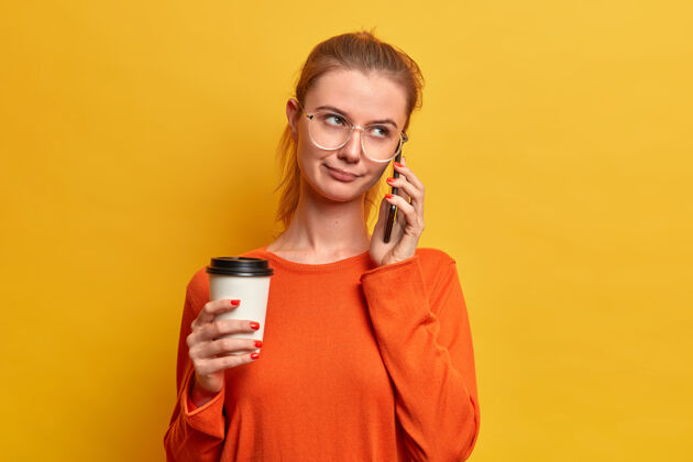 人严肃漂亮的欧洲女孩有无聊的电话交谈 闲逛 与人交谈 拿着一次性咖啡 使用现代科技 穿着毛衣 在黄色的墙上摆姿势漂亮电话眼镜