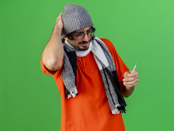 绿色疼痛的年轻白种人 戴着眼镜 戴着冬天的帽子 戴着围巾 手里拿着温度计 手放在头上 在绿色的背景上隔离 留着复印空间疾病手体温计