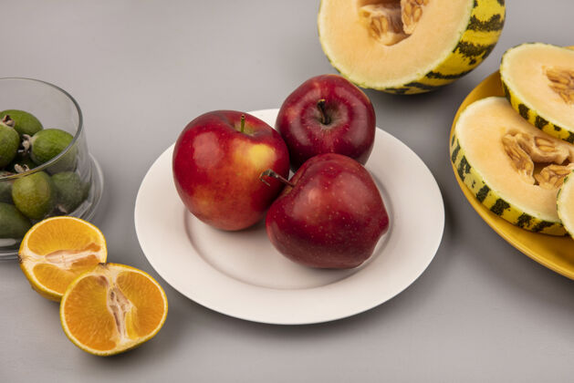 盘子白色盘子上的甜红苹果 黄色盘子上的蜜瓜片 灰色墙壁上的橘子被隔离 俯视图食品水果顶部