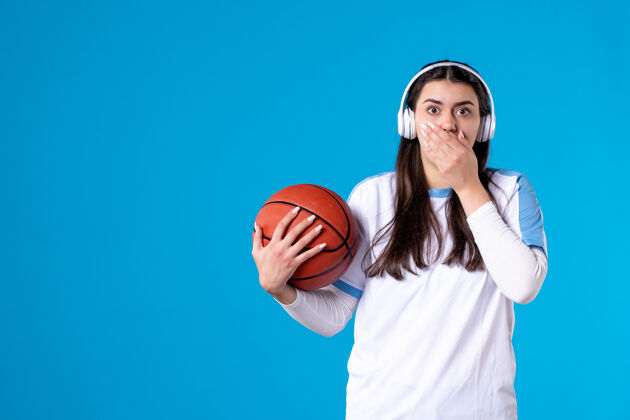 球正面图：戴着耳机拿着篮球的年轻女性站在蓝色的墙上体育比赛运动装