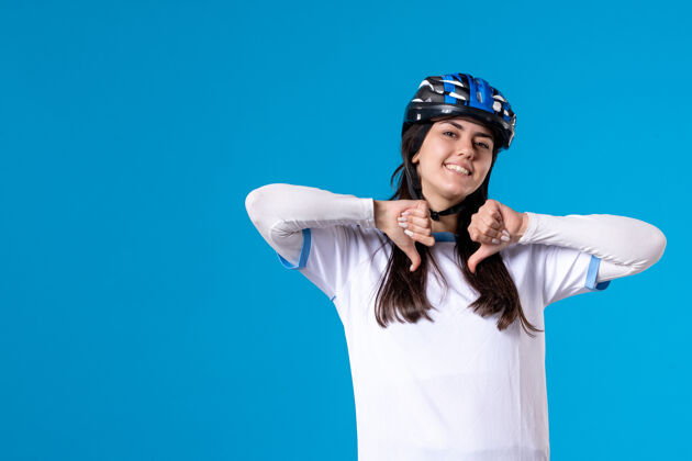 成人前视图穿着运动服的年轻女性 蓝色墙上戴着头盔拇指朝下年轻女性头盔