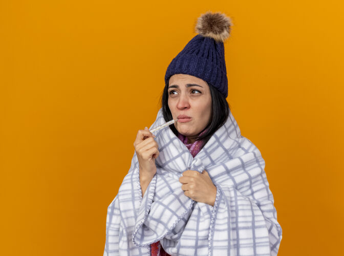 复制皱眉的年轻白种人生病的女孩戴着冬天的帽子 围巾裹着格子布 嘴里叼着温度计 抓着格子布 看着橙色背景上孤立的一面 还有复制空间拿着围巾空间