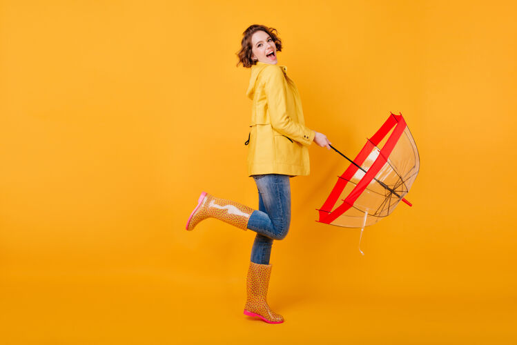 成人身着时尚秋季夹克的快乐女孩单腿站立的全幅照片兴奋的欧洲女模特打着伞在黄色墙上表达积极的情绪卷发天气明亮