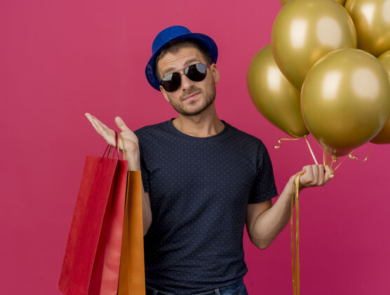 帽子戴着蓝色派对帽的自信帅哥 戴着太阳眼镜 手里拿着氦气球和纸购物袋 隔离在粉红色的墙上金气球市民