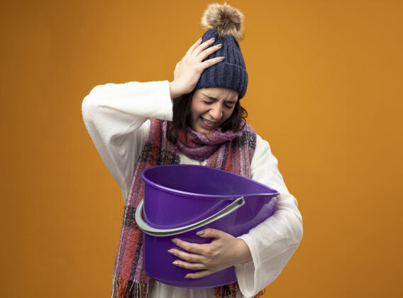 疾病疼痛的年轻生病的女人 戴着长袍 戴着冬天的帽子 戴着围巾 手里拿着塑料桶 恶心 手放在头上 闭着眼睛 隔离在橙色的墙上年轻人人姿势
