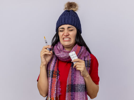 疾病恼怒的年轻生病的女人戴着冬天的帽子和围巾拿着注射器和药片包闭着眼睛隔离在白色的墙上关闭衣服包装