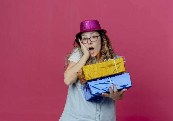 粉色惊讶的年轻女孩戴着眼镜和粉红色的帽子拿着礼盒 把手放在脸颊上孤立的粉红色眼镜惊喜盒子