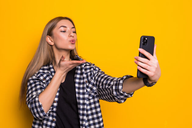 拍摄年轻漂亮的女人在电话里自拍吹吻隔离在黄墙上女性人物表演