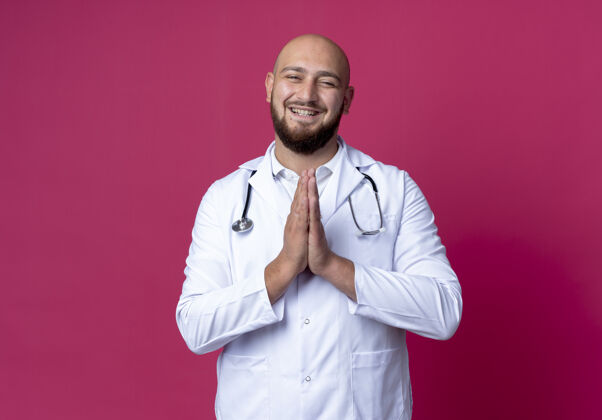 医疗面带微笑的年轻秃头男医生穿着医用长袍 听诊器显示出祈祷的手势隔离在粉红色祈祷粉色长袍
