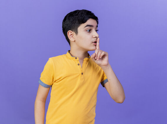 年轻人印象深刻的小男孩看着一边做沉默的手势孤立在紫色的墙上沉默姿势站立