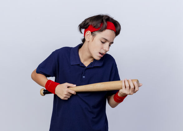 市民令人印象深刻的年轻英俊的运动男孩戴着头带和护腕与牙套举行和看棒球棒隔离在白墙上年轻男孩脸