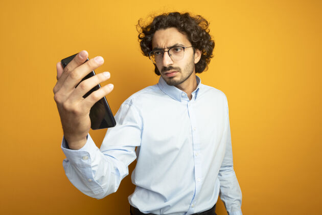 男人愁眉不展的年轻英俊的白人男子戴着眼镜拿着手机 看着橙色背景上孤立的有复印空间的手机眼镜
