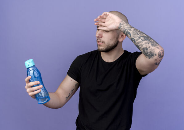 紫色年轻的运动型男人拿着水瓶 看着水瓶 把手腕放在紫色的额头上抱着年轻水