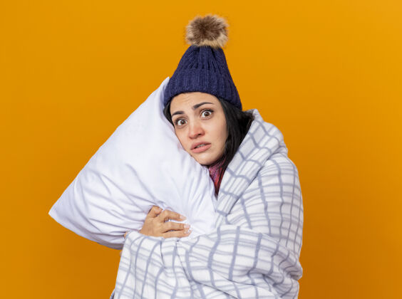 空间印象深刻的年轻白种人生病的女孩戴着冬天的帽子和围巾包裹在格子布抱枕看着相机孤立的橙色背景与复制空间女孩围巾印象
