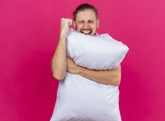 男人自信的年轻英俊的病夫抱着枕头望着前方挥舞着拳头尖叫着隔离在粉红色的墙上枕头拥抱人