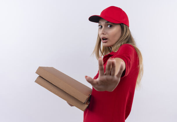 停止惊讶的年轻送货女孩穿着红色制服 戴着帽子拿着比萨饼盒 并显示停止手势孤立的白色制服女人手势