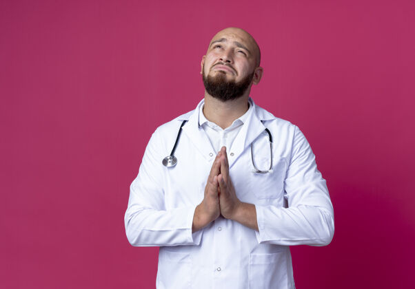 医生看着一位身穿医用长袍 戴着听诊器的年轻男性医生在粉红色的墙上孤零零地做着祈祷的姿势手势表演穿