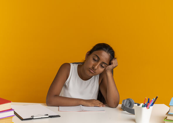 手疲惫的年轻女学生闭着眼睛坐在课桌旁 手里拿着学习用具 头放在桔子上桌子坐着桔子
