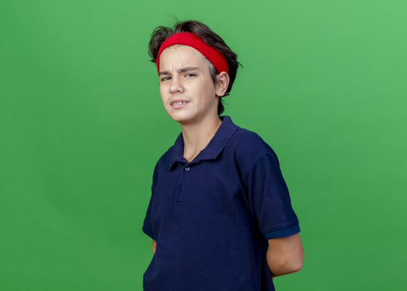 皱眉皱眉的年轻帅气的运动男孩戴着头带和护腕 戴着牙套 双手放在后面 看着绿色墙壁上孤立的前方衣服背带姿势