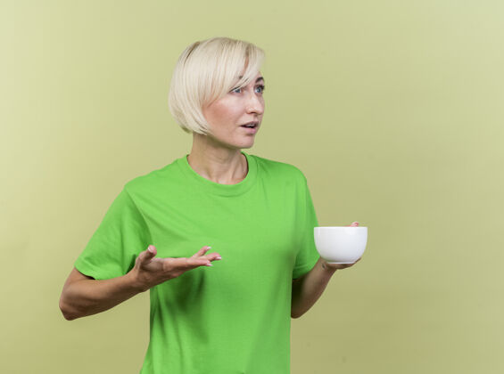 橄榄令人印象深刻的中年金发女人拿着一杯茶 在橄榄绿的墙上 一只空空的手看起来笔直孤立表情感情手
