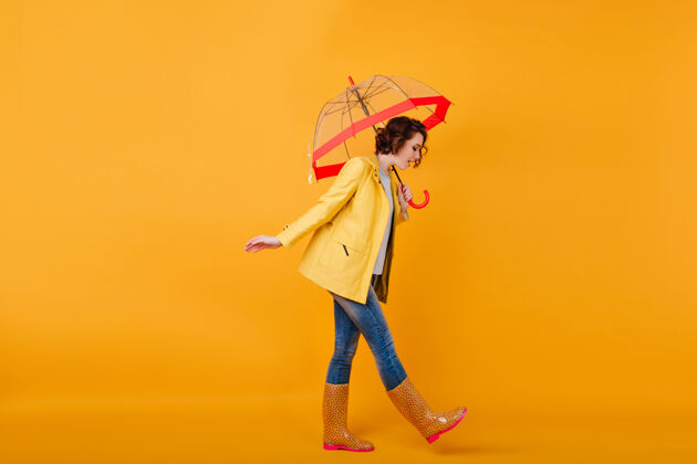 雨伞穿着胶鞋和黄色夹克的时髦女孩一边打着伞一边低头一个卷曲的短发女人穿着牛仔裤拿着阳伞走的摄影棚镜头表情阳伞年轻