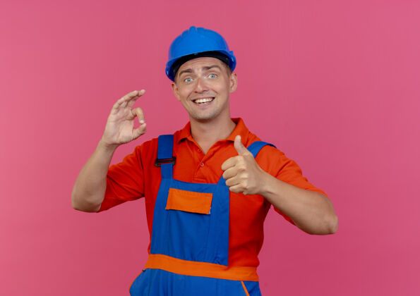 拇指年轻的男建筑工人穿着制服 戴着安全帽 在粉红色的地板上竖起大拇指手势头盔年轻