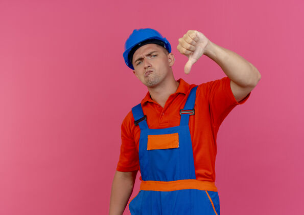 建筑工人穿着制服 戴着安全帽的年轻建筑工人把大拇指放在粉红色的地板上年轻向下男性
