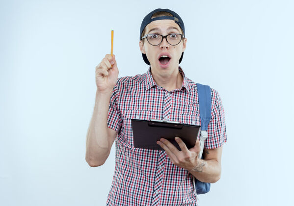 背景惊讶的年轻学生男孩背着书包 戴着眼镜 戴着帽子 拿着剪贴板 在白色的纸上举笔笔惊喜白色
