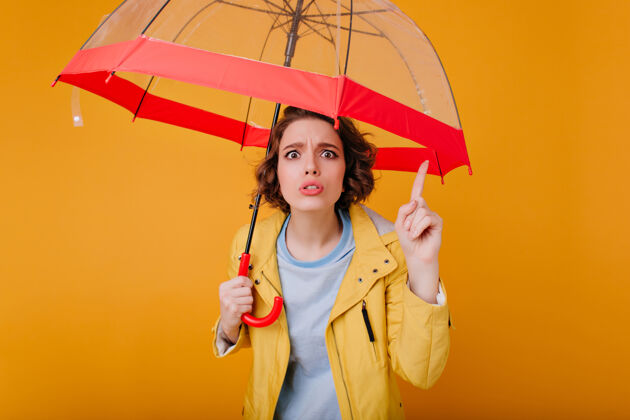 雨一个漂亮的女孩 波浪形的发型 一边打着时髦的雨伞 一边做着滑稽的表情一张不开心的穿着秋衣的白女人拿着红阳伞的照片明亮室内不快乐