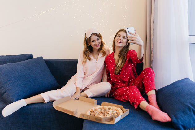 自拍可爱的卷曲的白袜子年轻女子微笑着她的朋友在家里自拍两个高兴的姐妹在周末吃比萨饼的室内照片披萨懒惰睡衣