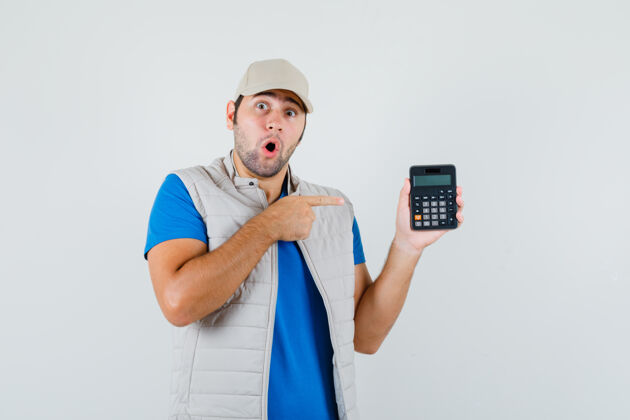 英俊一个穿着t恤衫 夹克衫 戴着帽子的年轻人指着计算器 惊讶地看着前方男人计算器夹克