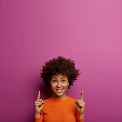 人类积极快乐的卷发女人获得新的发展理念 在上面点食指 在紫色的墙上展示复制空间 愉快地微笑 在室内摆姿势 为您展示美好的提议促销种族微笑