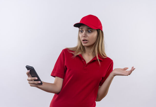 表情害怕的年轻送货女孩穿着红色制服 戴着帽子 看着手机上的白色隔离年轻电话帽子