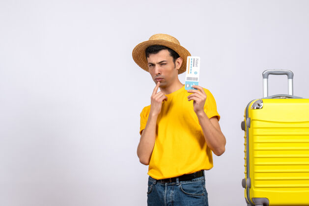 帽子前视图困惑的年轻游客穿着黄色t恤站在手提箱附近举着机票青少年手提箱T恤