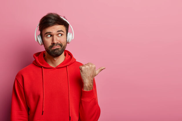 帅气好奇的大胡子男人的肖像在空白的右边指着拇指 戴着立体声耳机和红色休闲运动衫 展示了一些有趣的东西 孤立在粉色粉彩墙上站工作时尚