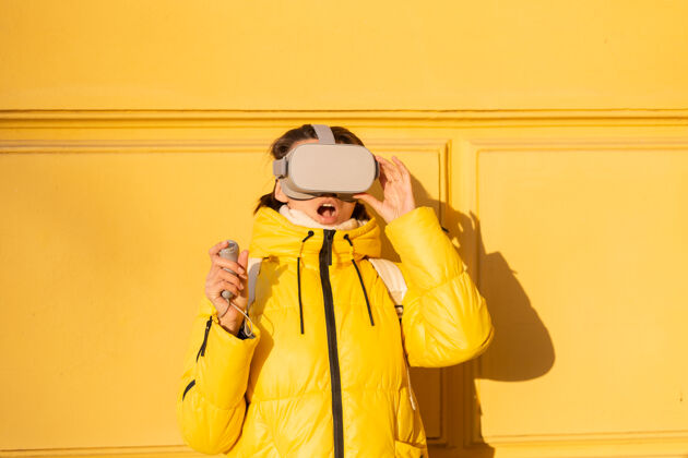 数码一个戴着虚拟现实眼镜的女人的肖像 在冬天的阳光下 穿着暖和的衣服 靠在黄色的墙上娱乐虚拟现实耳机女孩