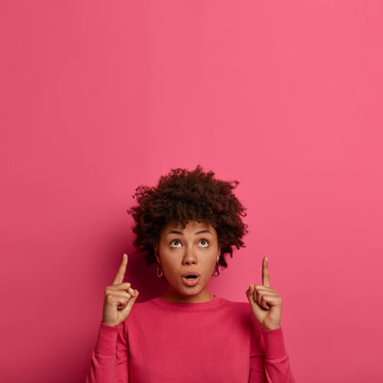 姿势尴尬震惊的非裔美国妇女的垂直镜头表示食指向上 令人印象深刻的东西 显示了粉红色墙上的空白 站在室内无语非洲介绍指向