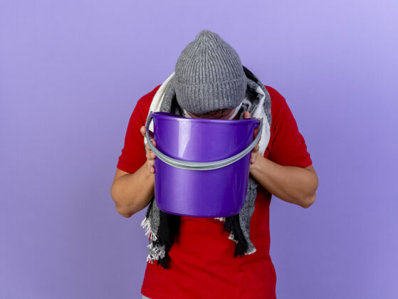姿势年轻英俊的金发病男子戴着冬天的帽子和围巾拿着塑料桶呕吐到它隔离在紫色的墙上人英俊感觉