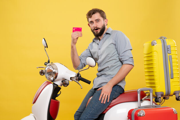 银行旅游概念的正面视图 一个留着胡须的年轻人坐在摩托车上 手提箱上挂着黄色的银行卡微笑成人年轻人