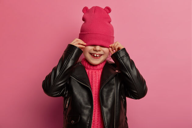 幼儿园积极的小朋友用粉色帽子遮住脸 遮住眼睛 穿皮夹克 有着俏皮快乐的微笑 对着玫瑰色的墙壁摆姿势 感觉乐观 试穿时髦的服装儿童概念人感觉积极