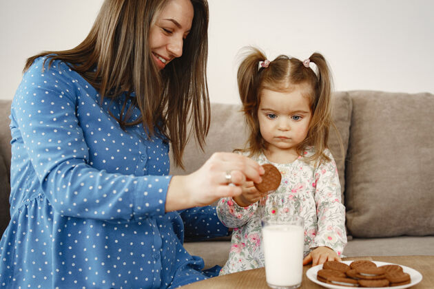 烘焙怀孕的妈妈穿着裙子女孩喝牛奶妈妈和女儿享受饼干给予女人快乐