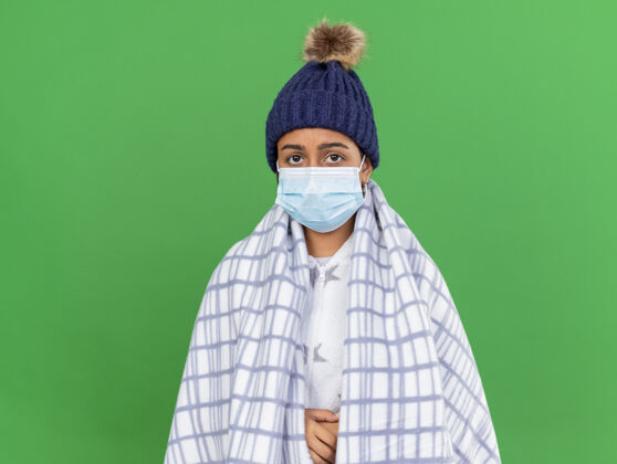 年轻年轻的生病女孩戴着冬天的帽子 戴着围巾和医用口罩 在绿色背景上用格子布包裹着面具格子围巾