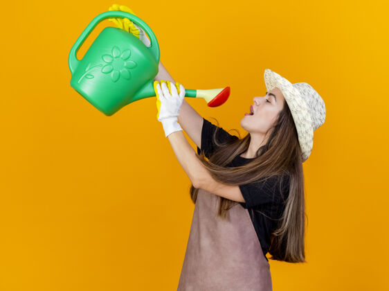 帽子很高兴美丽的园丁女孩穿着制服 戴着园艺帽 戴着手套 在橙色的背景上用浇水罐给自己浇水女孩手套她自己