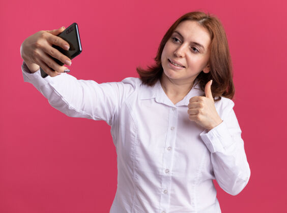 自拍身穿白衬衫的年轻女子手持智能手机自拍 微笑着竖起大拇指站在粉色的墙上站着抱着拇指