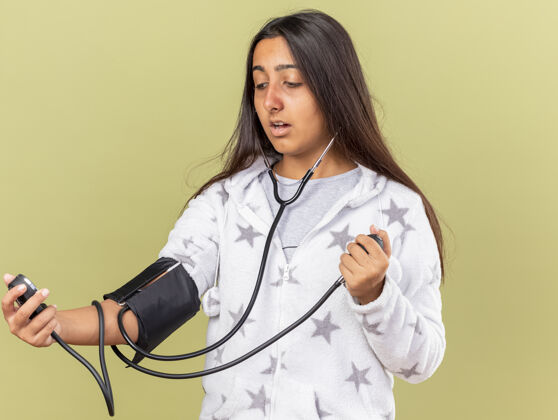 压力惊讶的年轻女孩用橄榄绿背景上的血压计测量自己的血压衡量疾病自己