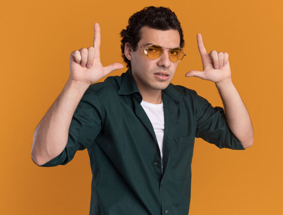 男人穿绿衬衫的年轻人戴着眼镜 自信地看着前面 用手指在橘色的墙上做着的手势衬衫表情自信