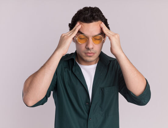 疲倦一个穿绿衬衫戴眼镜的年轻人站在白墙上摸着头累得头痛衬衫头年轻